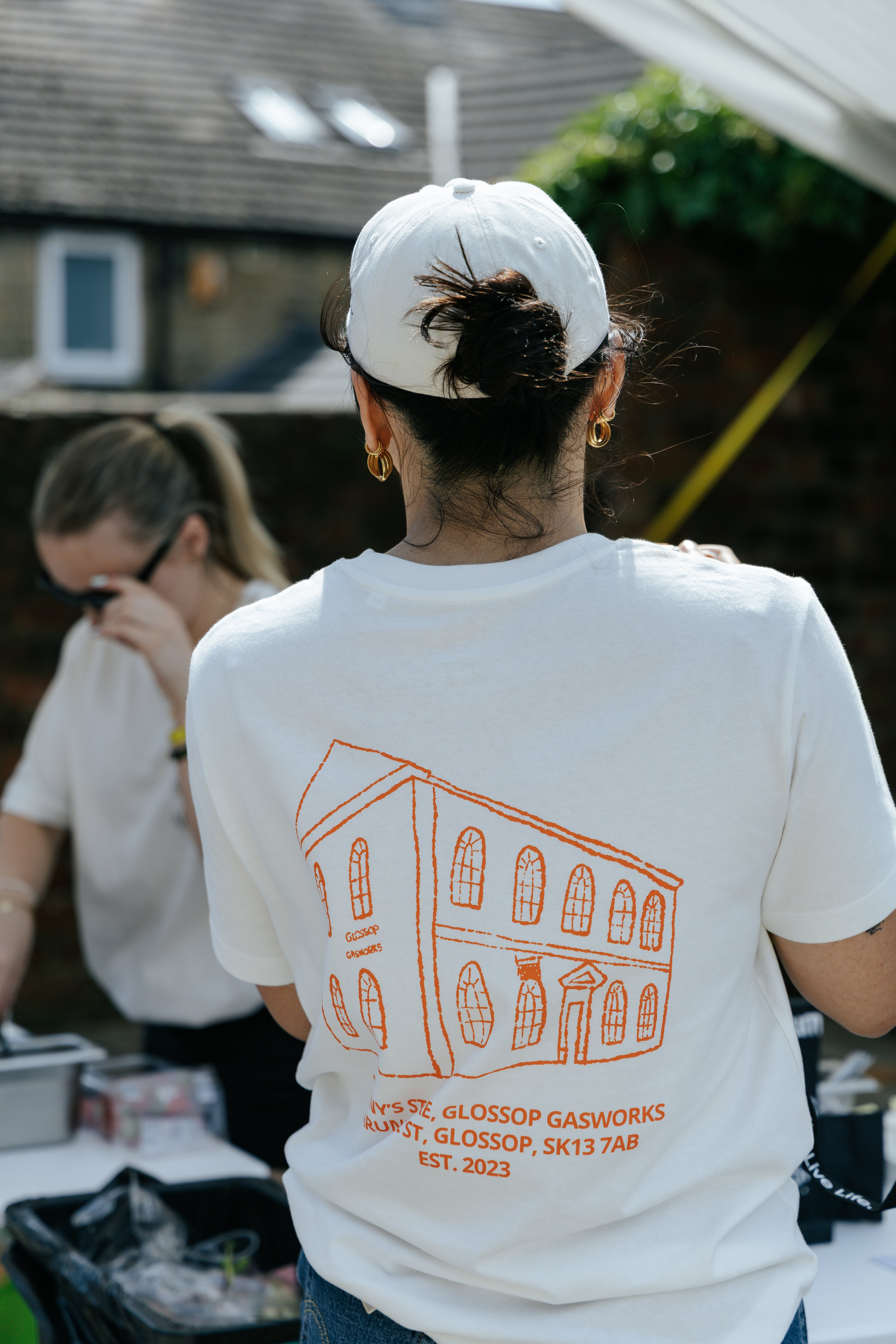 A person viewed from behind wearing a white t-shirt with an orange line drawing of a building and the text 'SUNNY'S STORE, GLOSSOP GASWORKS, ARUNDEL ST, GLOSSOP, SK13 7AB EST. 2023' printed on the back, along with a white baseball cap in an outdoor setting.