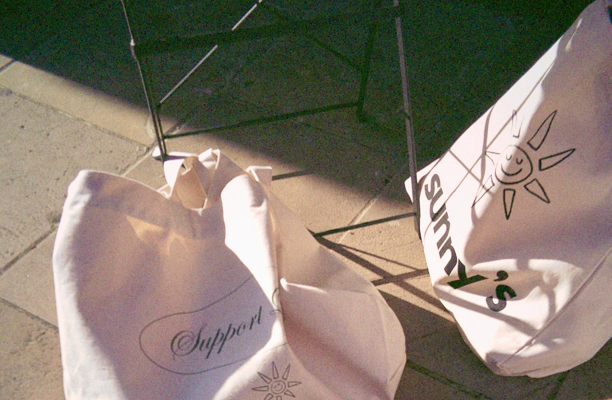Two off-white canvas tote bags with black printed graphics and text, resting on a stone pavement under harsh direct sunlight. 