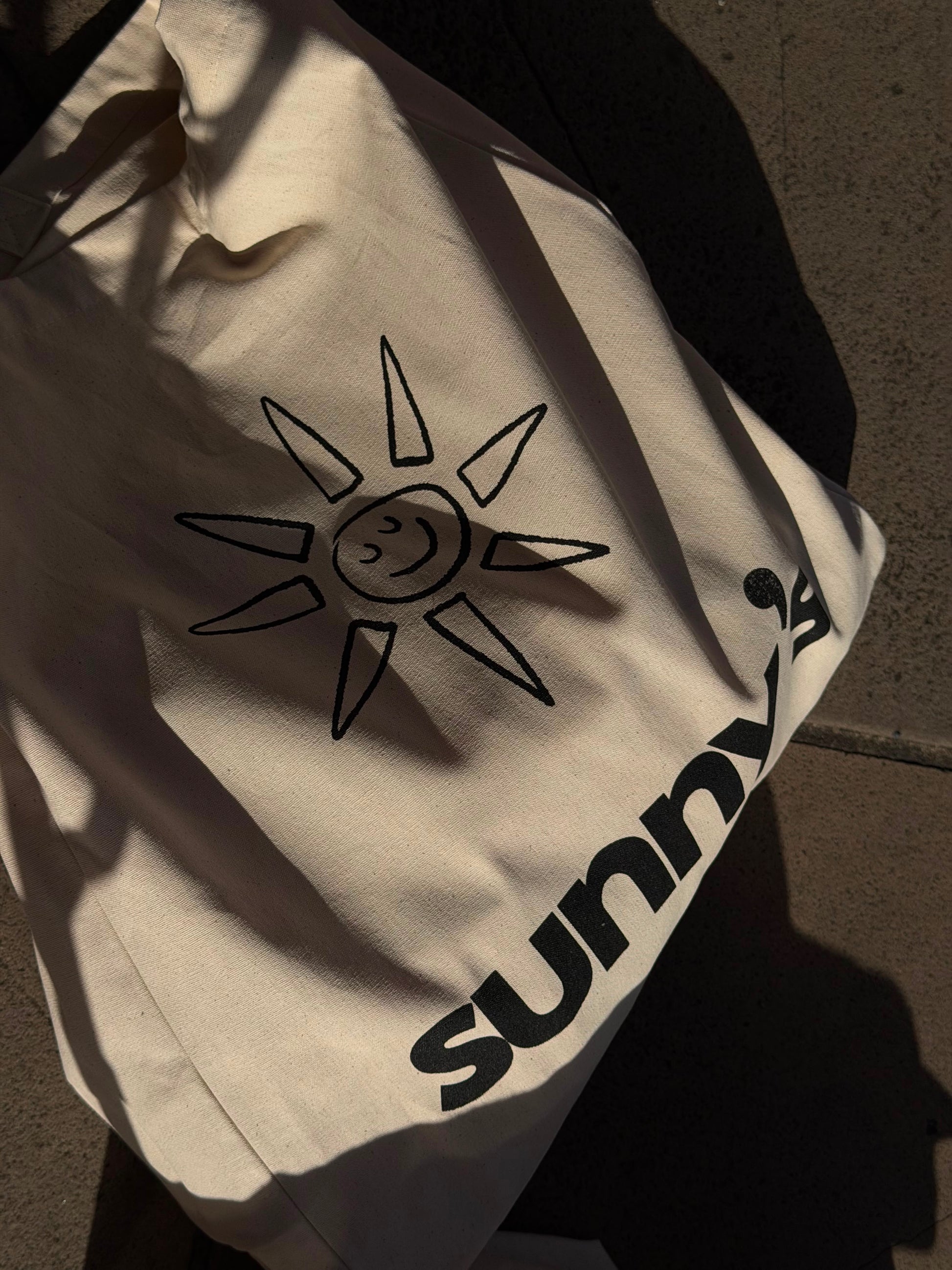A cream-colored canvas tote bag with a black hand-drawn sun graphic and the 'sunny's' printed in black, shown in dramatic natural sunlight on concrete.