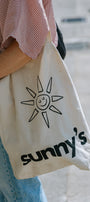 A person wearing a red-and-white striped shirt and blue jeans carries a cream canvas tote bag with a black sun graphic and the text 'sunny's' printed on the front.