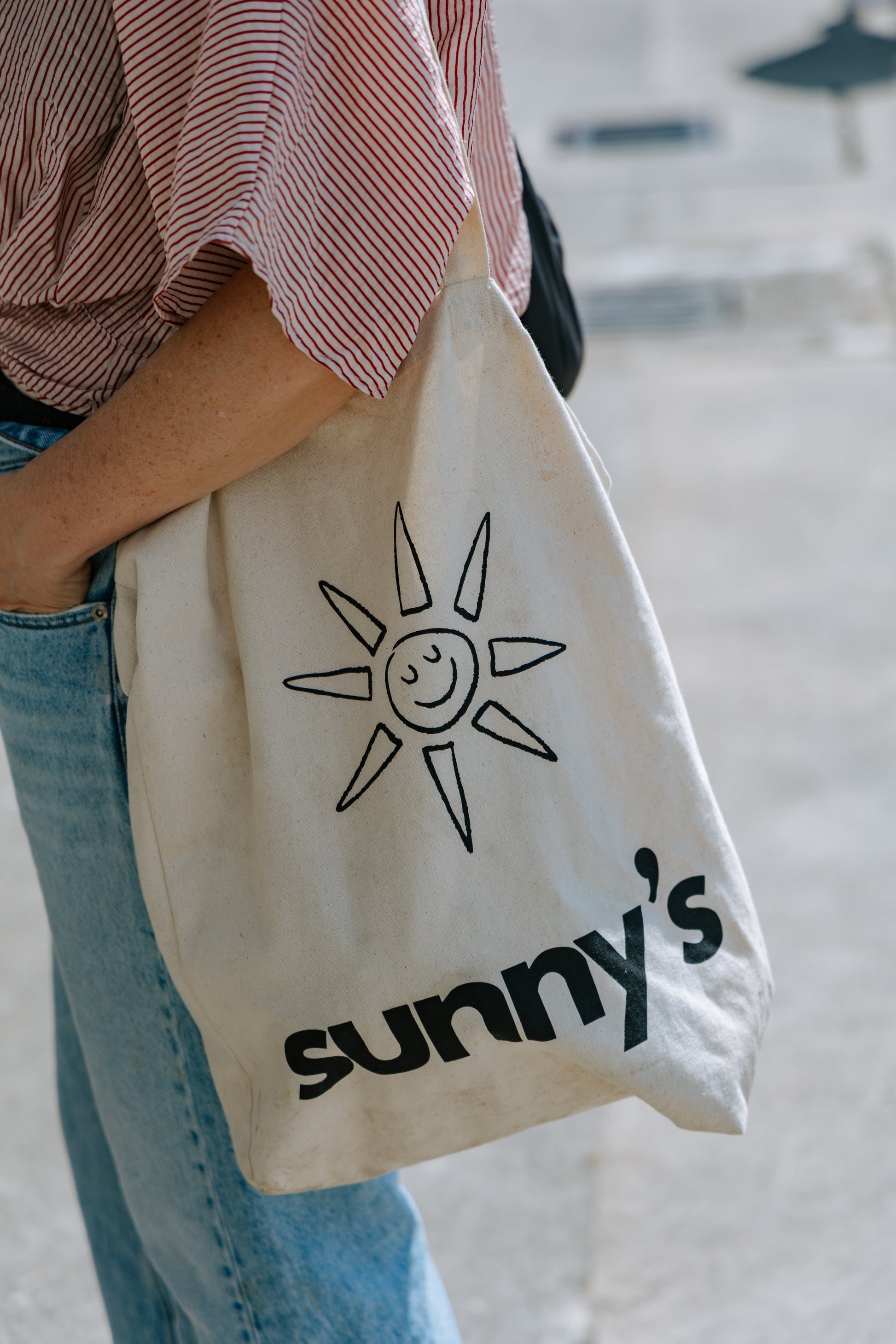 A person wearing a red-and-white striped shirt and blue jeans carries a cream canvas tote bag with a black sun graphic and the text 'sunny's' printed on the front.