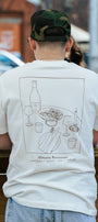 A person viewed from behind wearing a white graphic t-shirt with a dark brown line art illustration of a restaurant table scene and the text 'Alimentae Restaurant'.