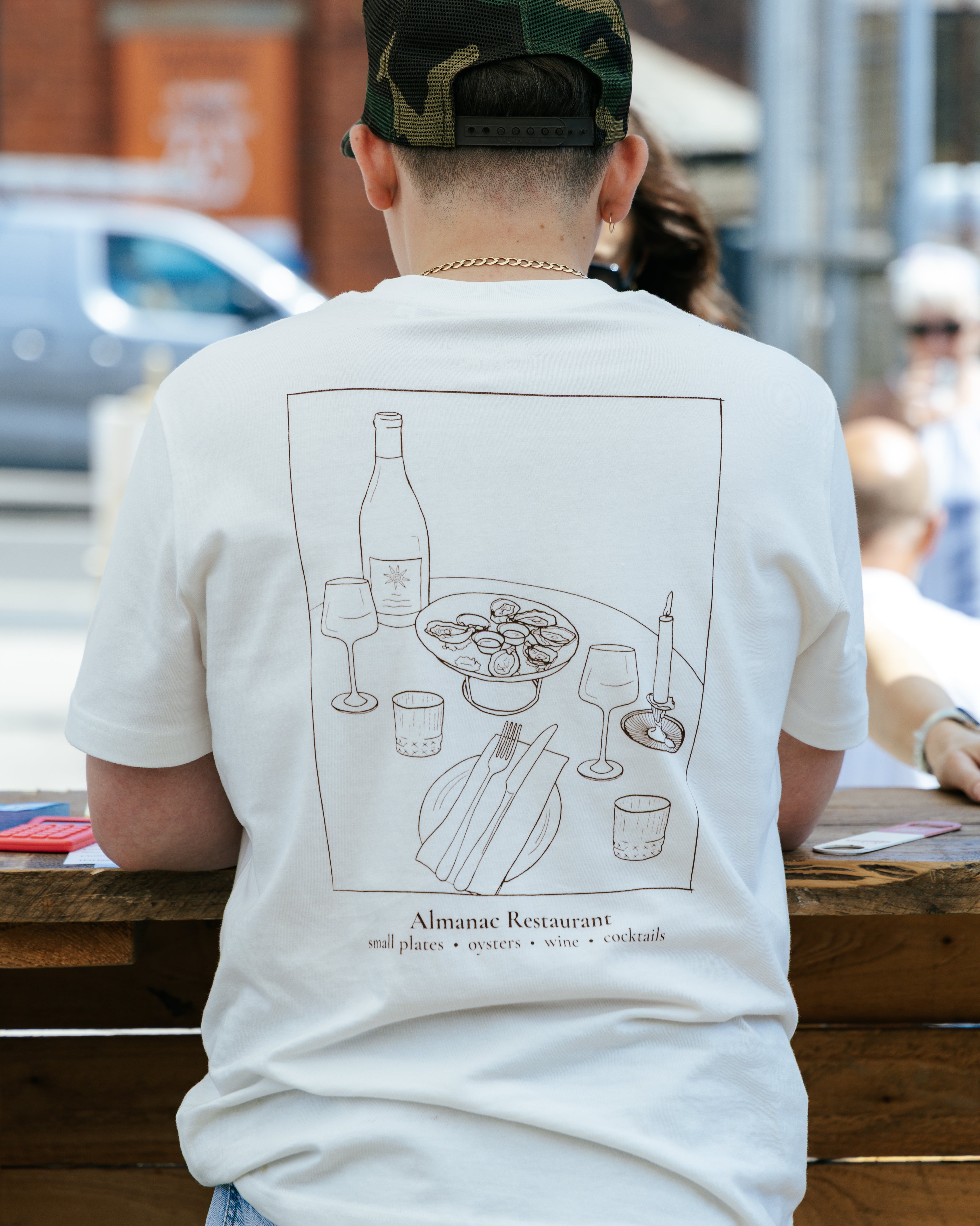 A person viewed from behind wearing a white graphic t-shirt with a dark brown line art illustration of a restaurant table scene and the text 'Alimentae Restaurant'.