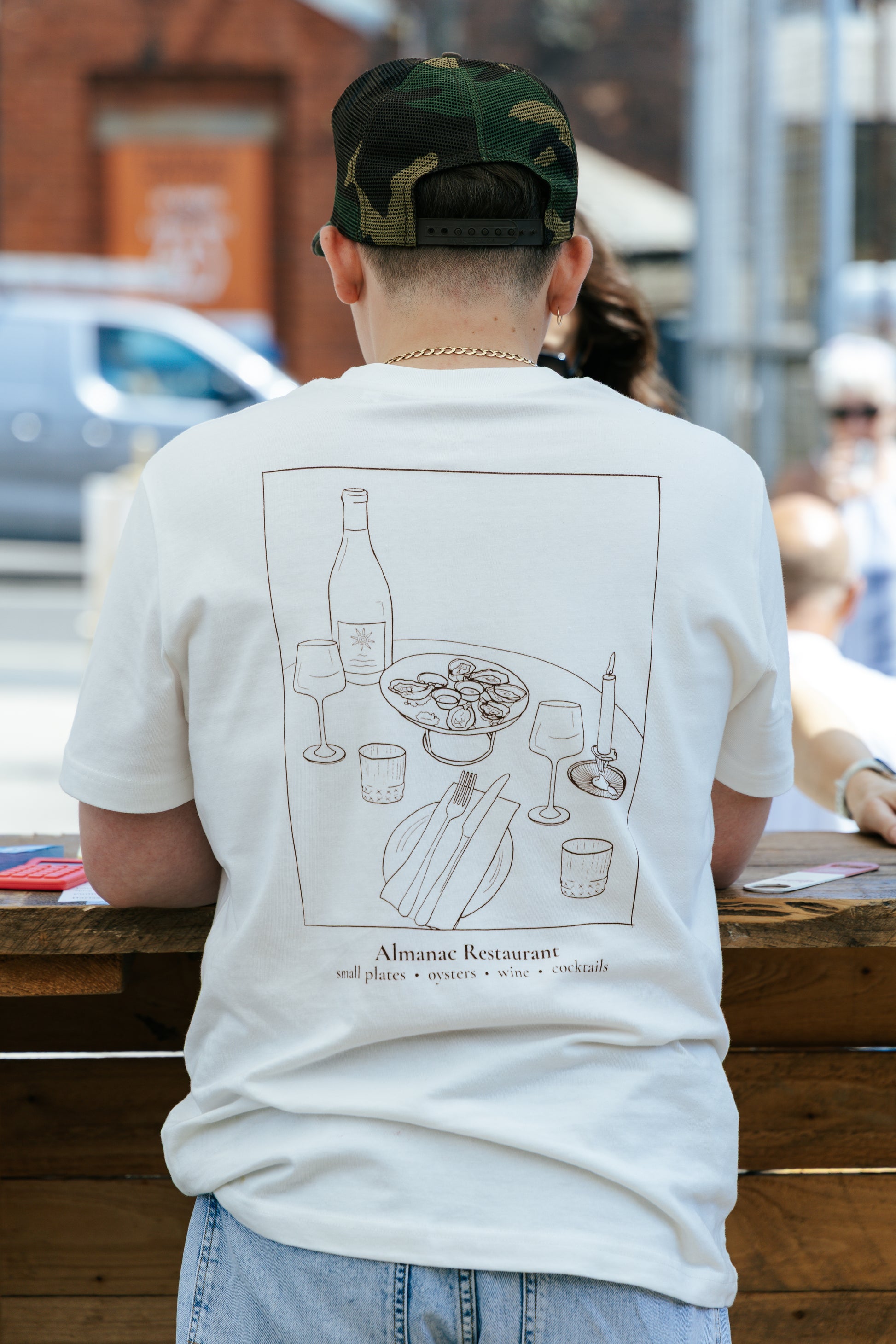 A person viewed from behind wearing a white graphic t-shirt with a dark brown line art illustration of a restaurant table scene and the text 'Alimentae Restaurant'.