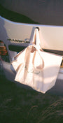 A cream canvas tote bag with black script text hanging from a hook on the back of a white van in a dark outdoor setting.