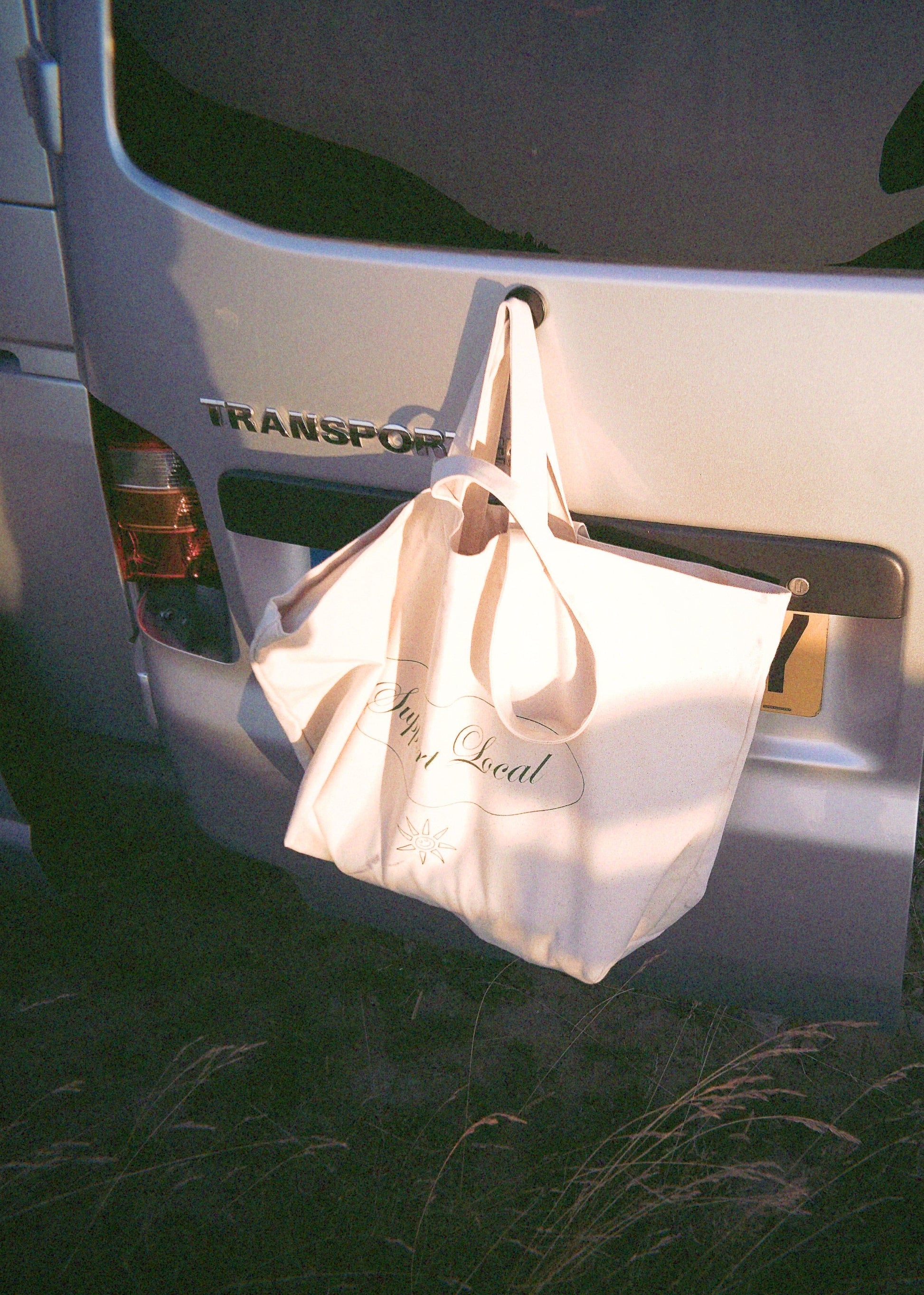 A cream canvas tote bag with black script text hanging from a hook on the back of a white van in a dark outdoor setting.