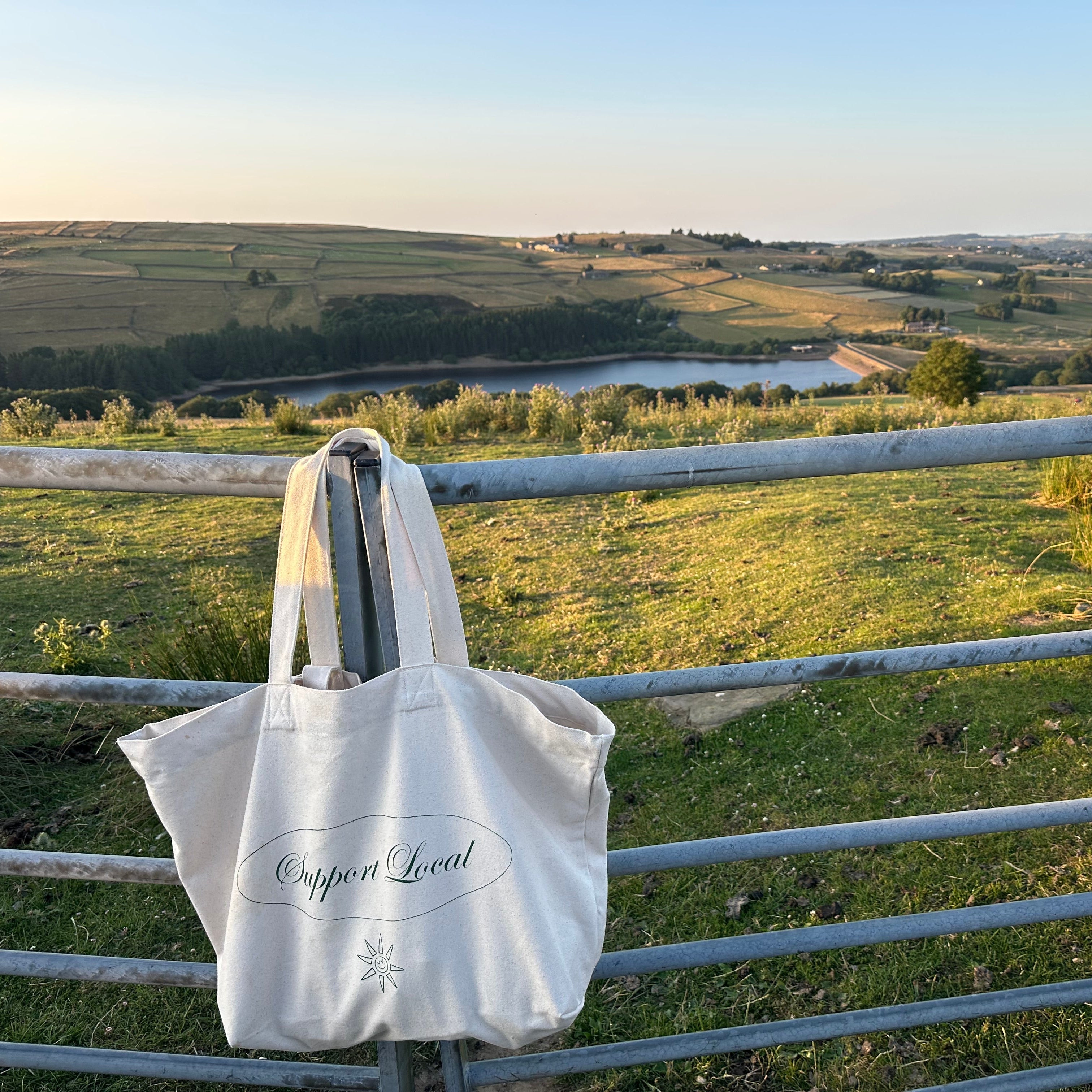 A white canvas tote bag with 'Support Local' printed on the front, hanging from a metal fence in a grassy field with a scenic rural landscape and a lake in the background.
