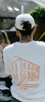 A person viewed from behind wearing a white t-shirt with an orange line drawing of a building and the text 'SUNNY'S STORE, GLOSSOP GASWORKS, ARUNDEL ST, GLOSSOP, SK13 7AB EST. 2023' printed on the back, along with a white baseball cap in an outdoor setting.