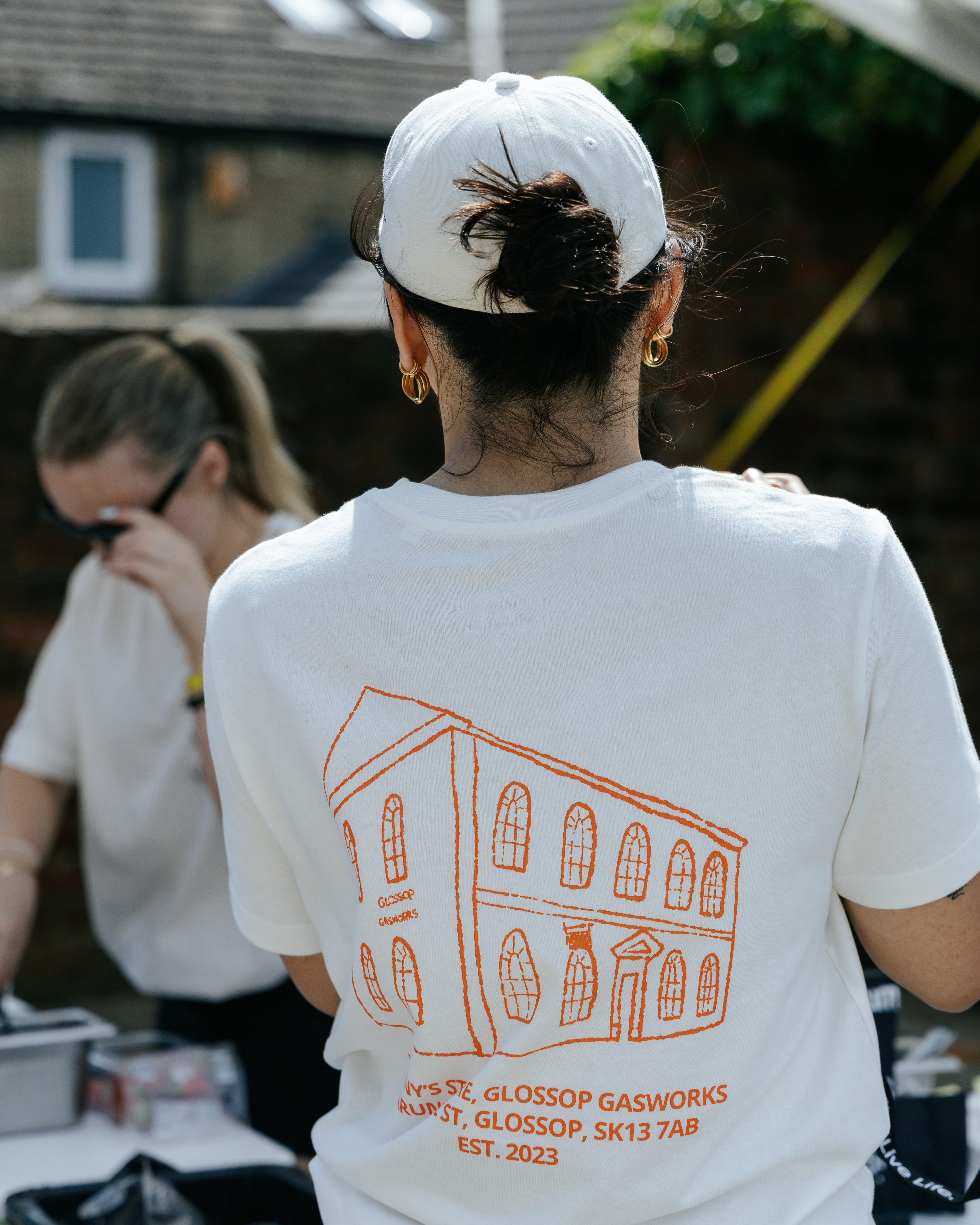 A person viewed from behind wearing a white t-shirt with an orange line drawing of a building and the text 'SUNNY'S STORE, GLOSSOP GASWORKS, ARUNDEL ST, GLOSSOP, SK13 7AB EST. 2023' printed on the back, along with a white baseball cap in an outdoor setting.