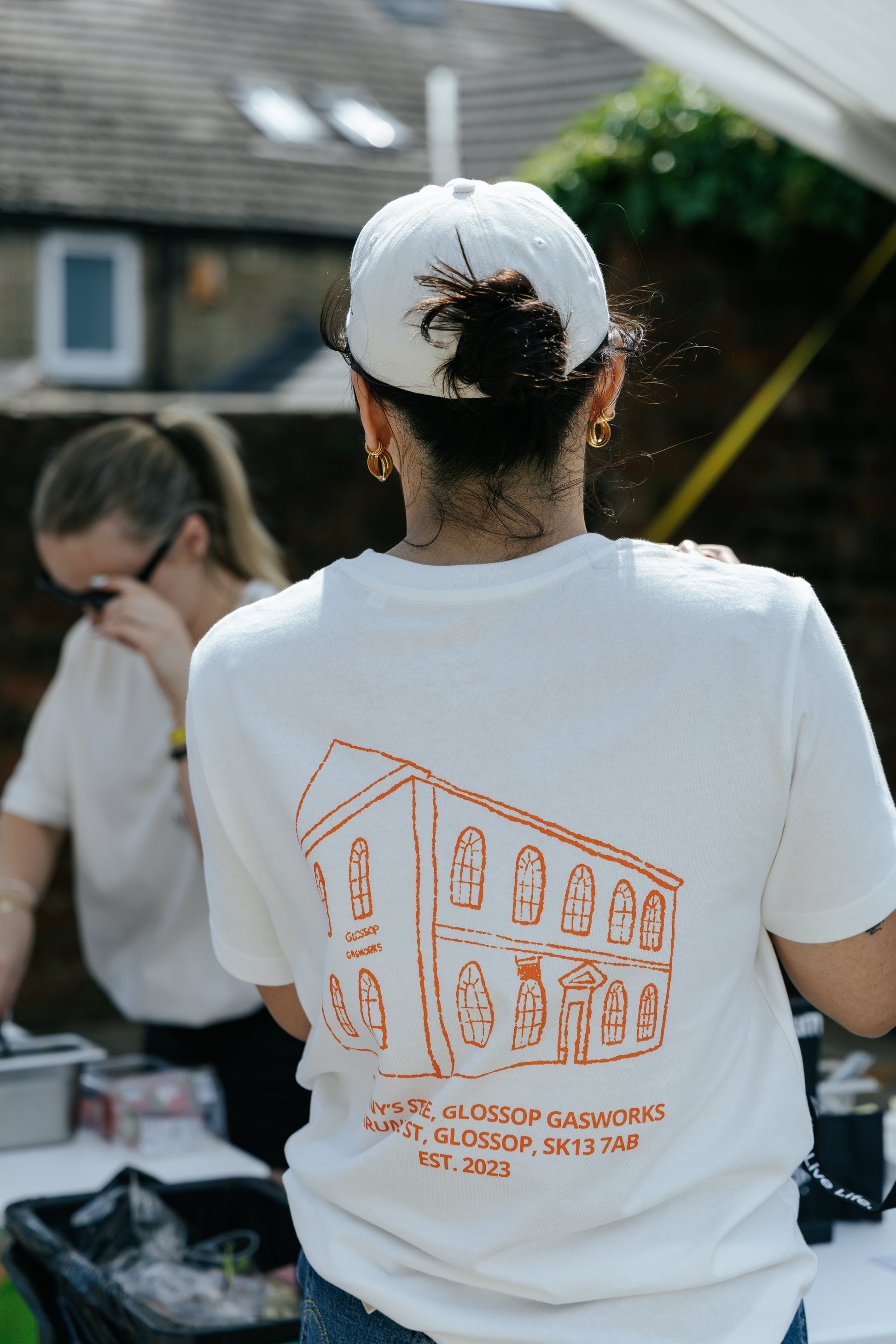 A person viewed from behind wearing a white t-shirt with an orange line drawing of a building and the text 'SUNNY'S STORE, GLOSSOP GASWORKS, ARUNDEL ST, GLOSSOP, SK13 7AB EST. 2023' printed on the back, along with a white baseball cap in an outdoor setting.