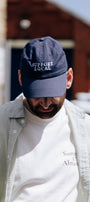 A man wearing a navy blue baseball cap with 'SUPPORT LOCAL' embroidered in white on the front, layered over a white t-shirt and a light-colored button-up shirt, photographed outdoors with a blurred brick building in the background.