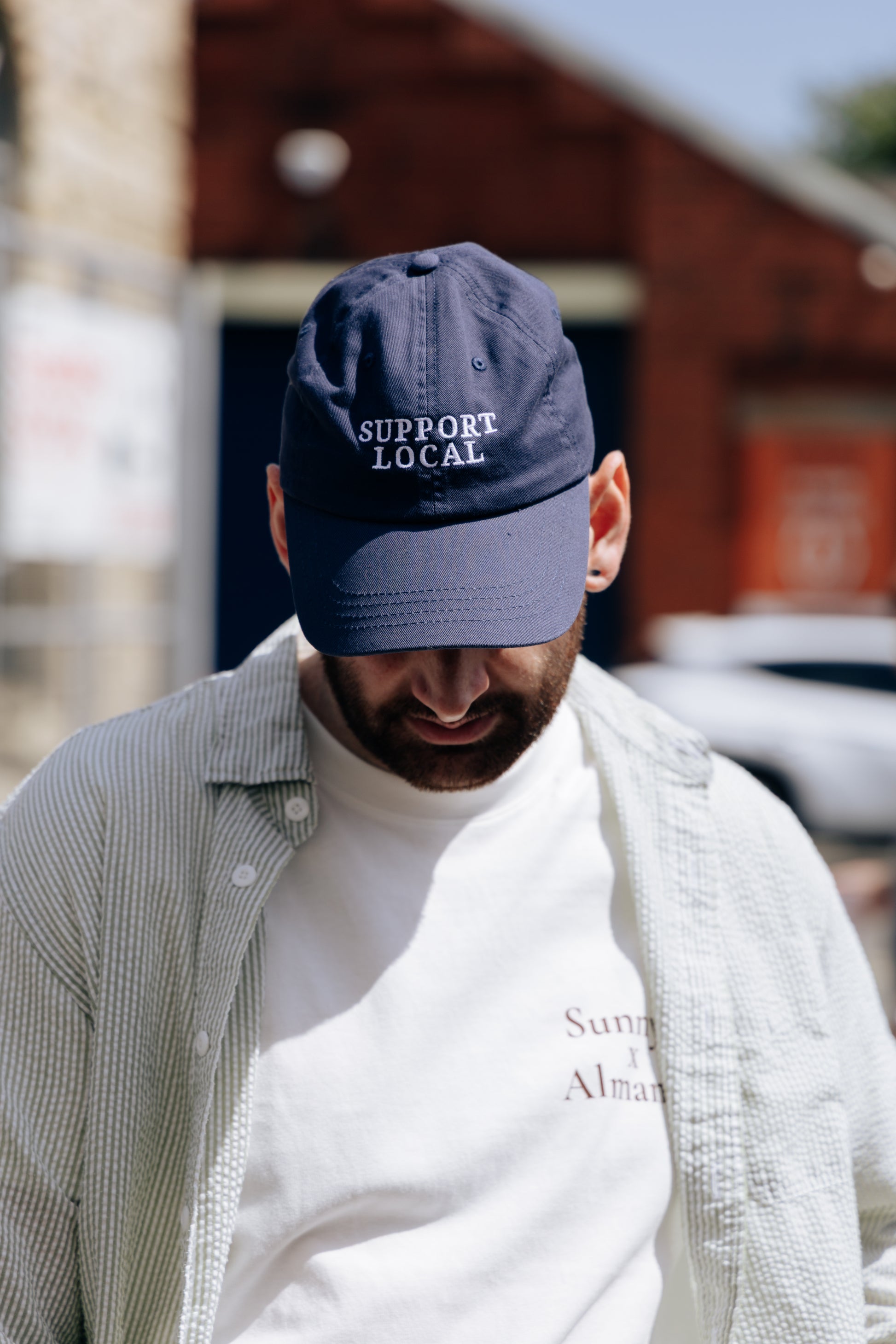 A man wearing a navy blue baseball cap with 'SUPPORT LOCAL' embroidered in white on the front, layered over a white t-shirt and a light-colored button-up shirt, photographed outdoors with a blurred brick building in the background.