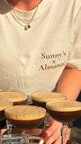 A person wearing a cream-colored t-shirt with 'Sunny's x Almanac' printed on the front, holding a silver tray with five espresso martinis in coupe glasses.