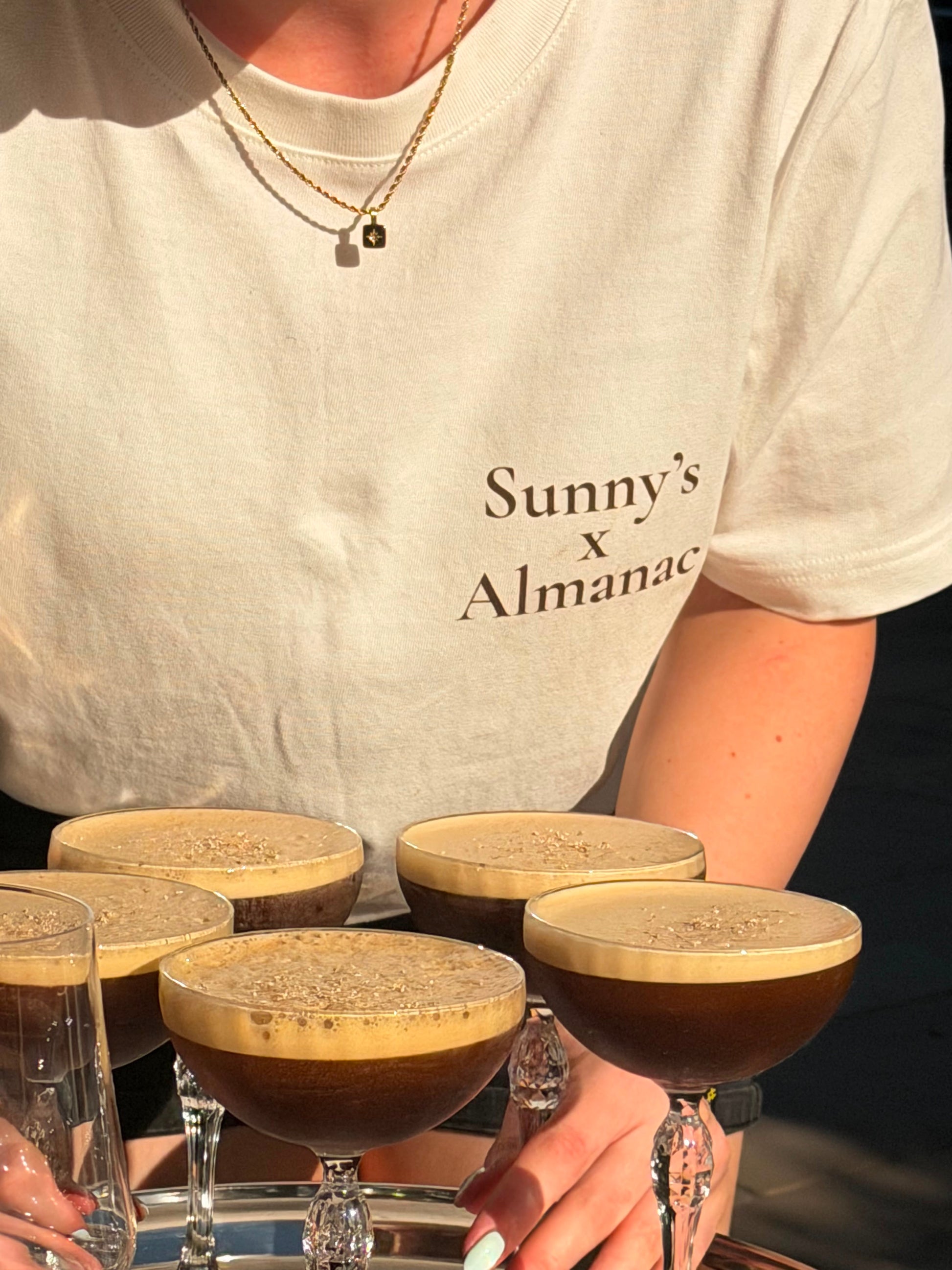 A person wearing a cream-colored t-shirt with 'Sunny's x Almanac' printed on the front, holding a silver tray with five espresso martinis in coupe glasses.