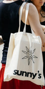 A cream canvas tote bag with a black sun graphic and the text 'sunny's' worn over the shoulder of a person in a black tank top and red shorts, standing outdoors next to a woman in a black outfit.