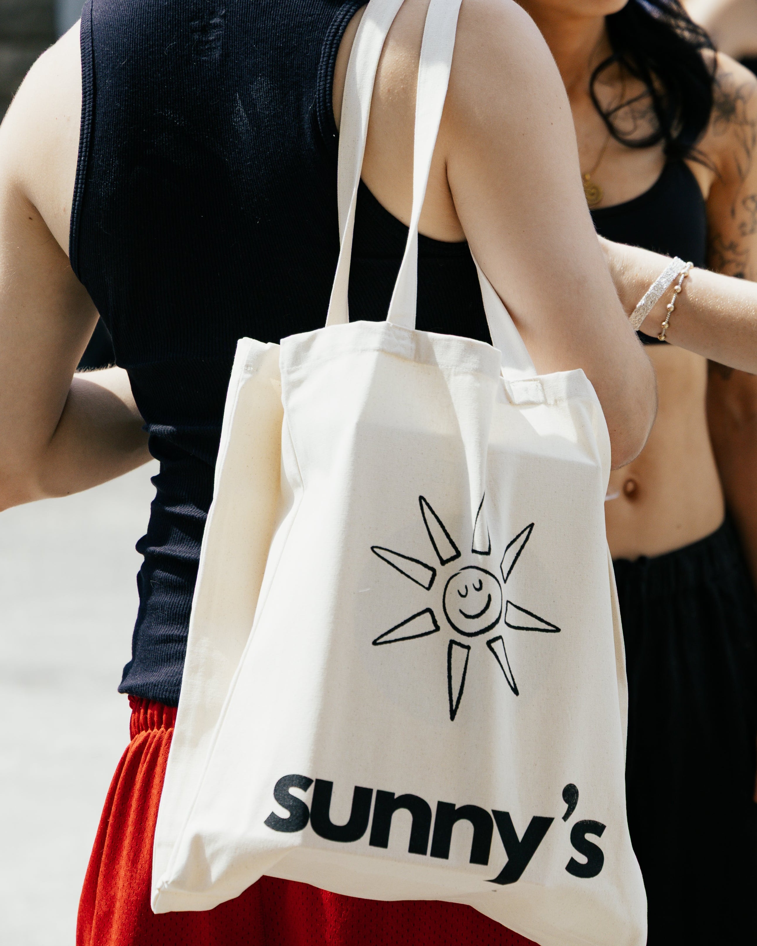 A cream canvas tote bag with a black sun graphic and the text 'sunny's' worn over the shoulder of a person in a black tank top and red shorts, standing outdoors next to a woman in a black outfit.