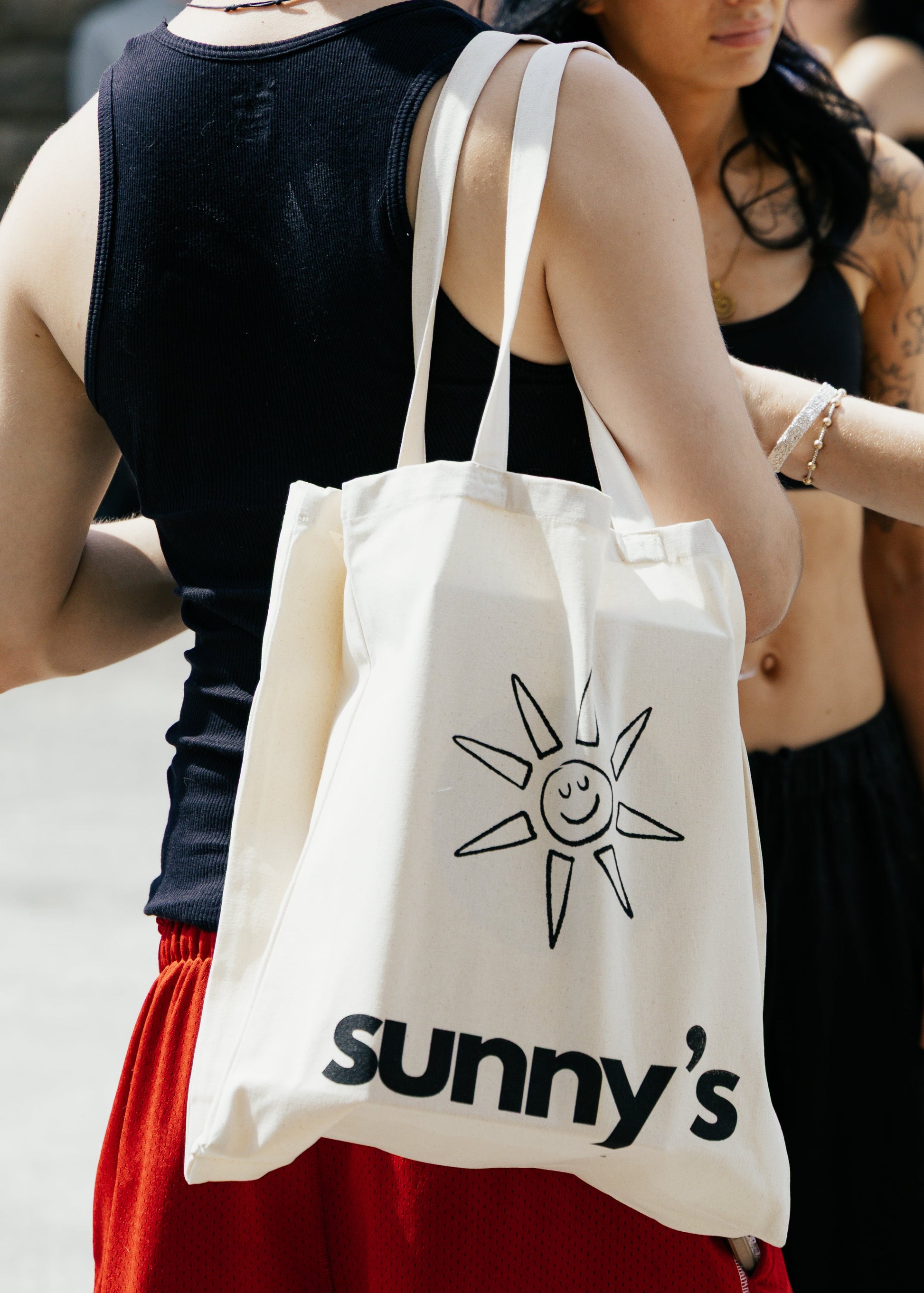 A cream canvas tote bag with a black sun graphic and the text 'sunny's' worn over the shoulder of a person in a black tank top and red shorts, standing outdoors next to a woman in a black outfit.