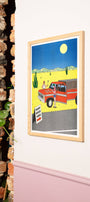 Framed artwork of a red truck in a desert scene on a white wall next to a brick wall.