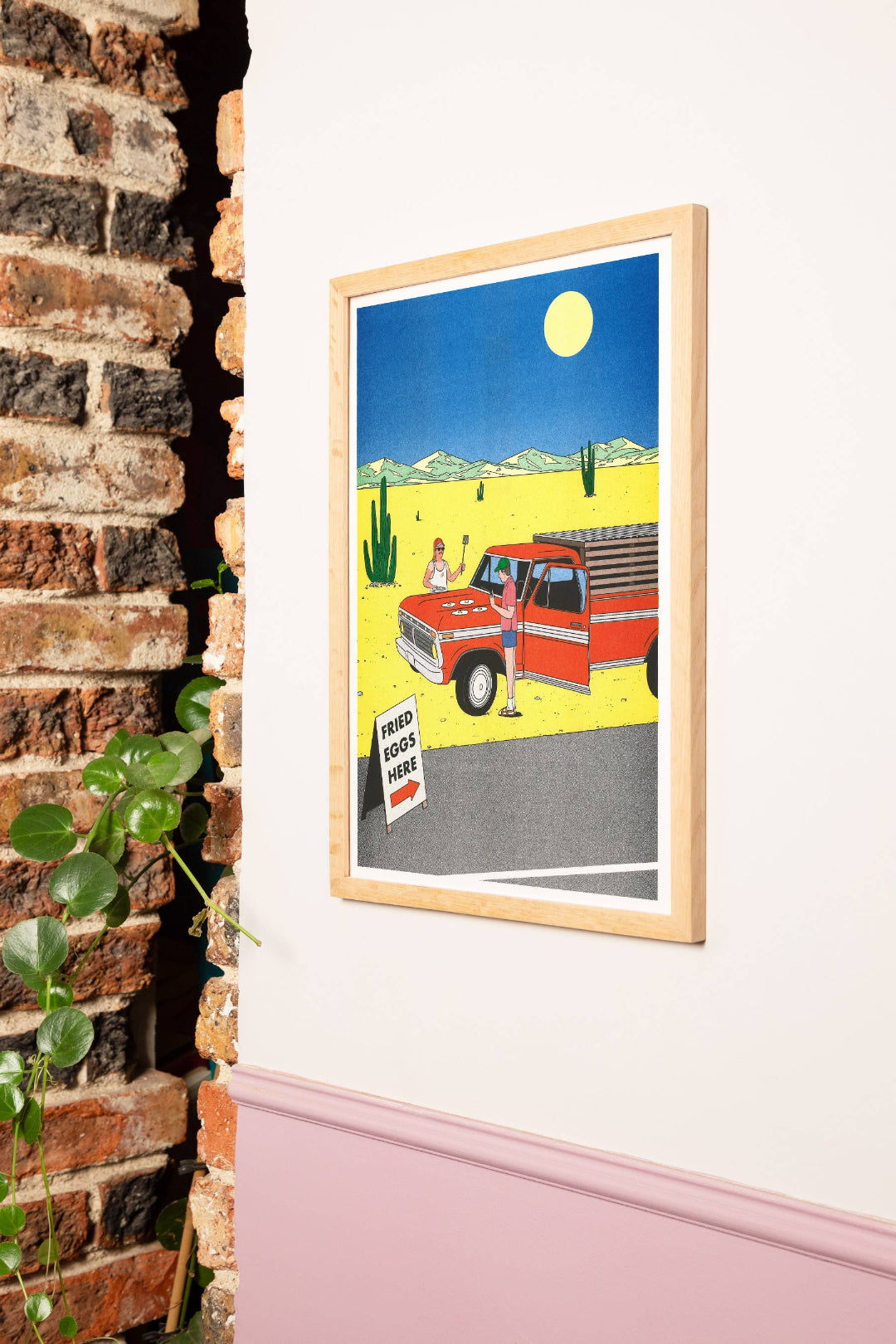 Framed artwork of a red truck in a desert scene on a white wall next to a brick wall.